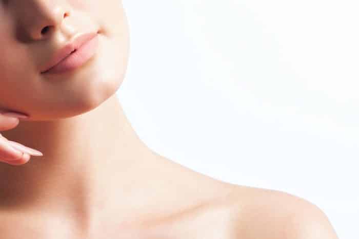 A close-up image of a person's lower face and neck with smooth, fair skin. The head is slightly tilted, and fingers lightly touch the chin. The background is a soft gradient of white, emphasizing the clear and clean complexion.