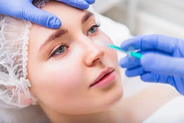 A person with a white hair cap receives a facial injection from a professional wearing blue gloves. The recipient's eyes are open, and they appear calm and relaxed. The setting suggests a medical or cosmetic procedure environment.