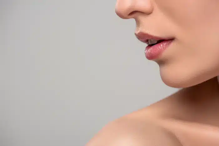 Close-up of a woman's lower face and neck against a plain background. The image focuses on her smooth skin, slightly parted lips, and part of her shoulder. She appears to be wearing light makeup, emphasizing her natural features.
