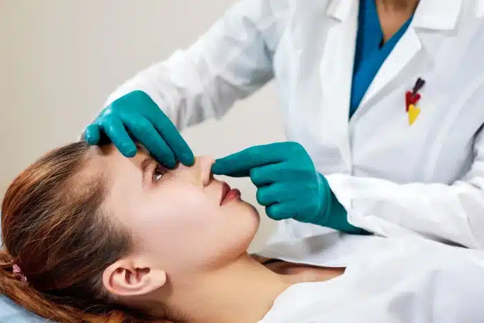 A woman lies down while a healthcare professional in a white coat and teal gloves examines her nose by gently pressing and adjusting it.