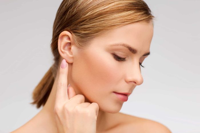 A woman with smooth skin and blonde hair tied back in a ponytail touches her ear with her finger. She is looking down with her eyes closed and has a serene expression. The background is plain and light-colored.