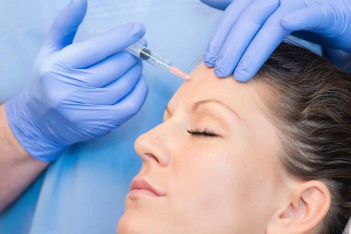 A person with closed eyes is receiving a facial injection. A medical professional wearing blue gloves administers the injection on the person's forehead. The background is a blur of blue, indicating a clinical setting.