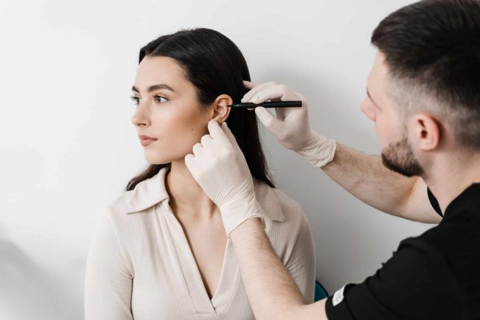 A person with dark hair, wearing a light-colored top, sits while another person in a dark shirt and wearing gloves marks their ear with a pen, possibly for a medical or cosmetic procedure, against a plain white background.