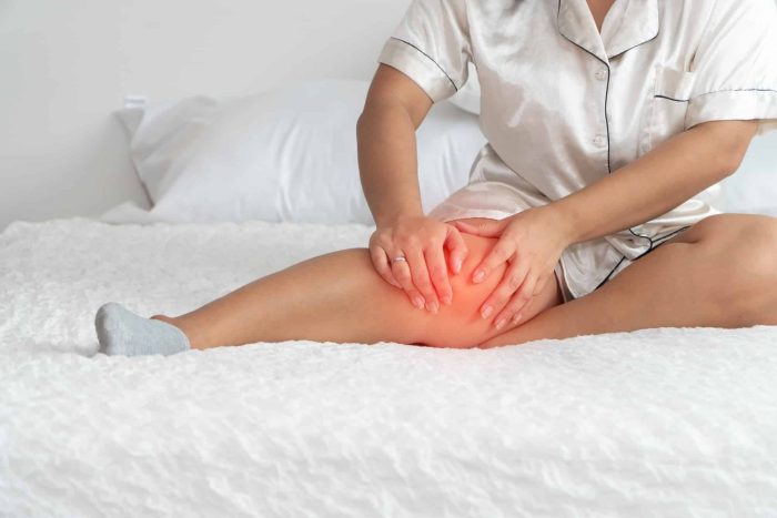 Person sitting on a bed, wearing pajamas and one sock, holding their knee which appears red, indicating pain or discomfort. The background shows a white pillow and bedding.