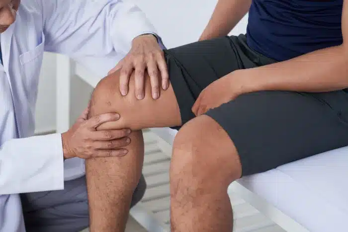 A doctor in a white coat examines the knee of a patient wearing black shorts and sitting on an examination table. The doctor's hands are on either side of the patient's knee, indicating a physical examination.