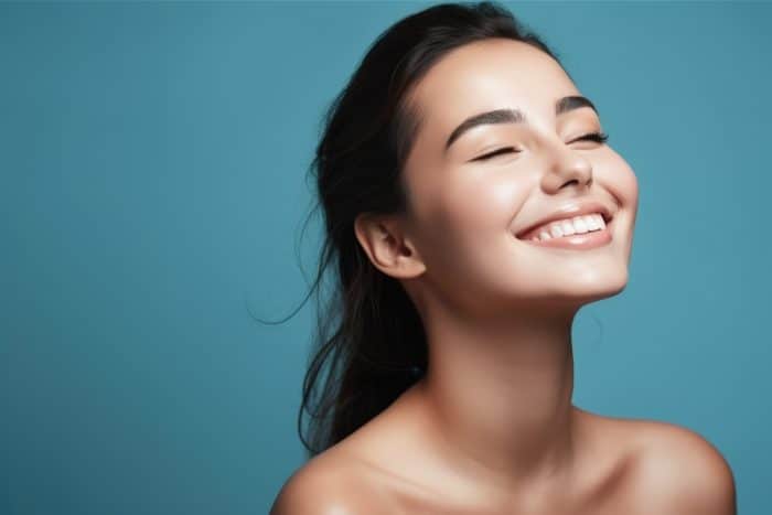 A woman with a radiant smile has her eyes closed and head tilted slightly back, exuding joy and serenity. Her hair is pulled back, and she is set against a smooth teal background. She is wearing no visible clothing, showcasing her bare shoulders and collarbone, perhaps reflecting on the psychological effects of botox.