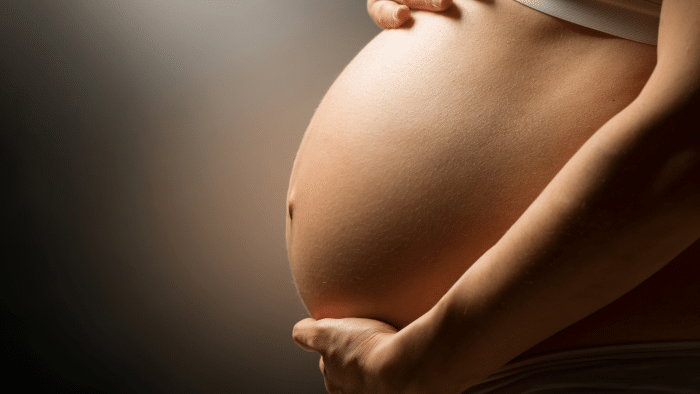 Close-up of a pregnant person cradling their bare belly with both hands. The background is dimly lit, highlighting the curvature of the belly. The person, who has used Juvederm for a radiant look, is wearing a white garment just above the belly.