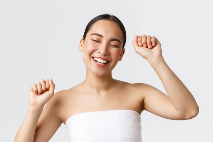A woman with straight dark hair is smiling with her eyes closed, the joy evident in her expression as she raises her arms. Clad in a white towel wrapped around her body, she radiates happiness. The plain light background makes her serene moment even more pronounced, unburdened by concerns like Dysport for bunny lines.