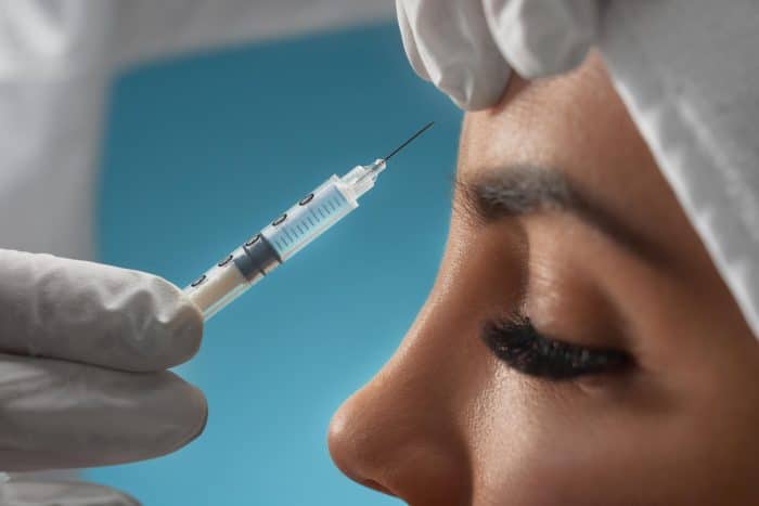 Close-up of a person's face with their eyes closed, wearing a white headcover. A gloved hand is holding a syringe, injecting Dysport into the forehead. Despite the potential for minor bruising, the scene appears calm against a blue background.