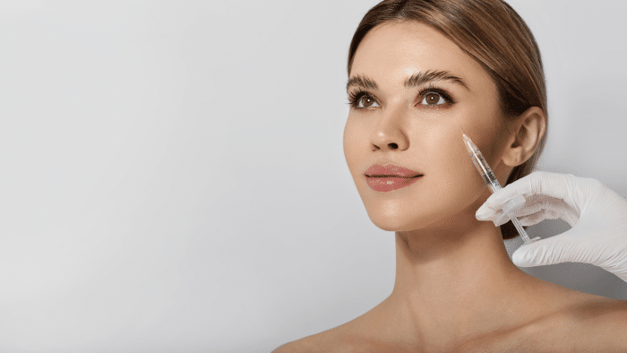 A woman with light brown hair and bare shoulders looks upwards while a gloved hand holds a syringe near her cheek, indicating a facial cosmetic procedure with Restylane vs Dysport. The background is light grey.