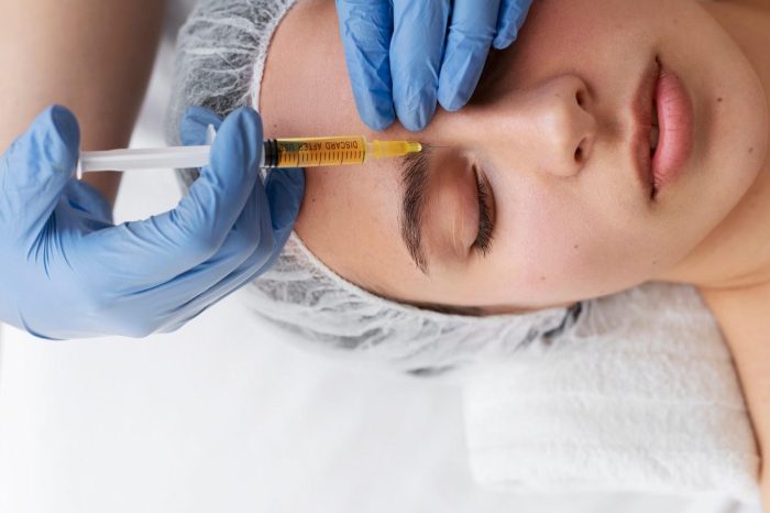 A person receiving a facial injection of Dysport between the eyebrows from a clinician wearing blue gloves. The individual, lying down with eyes closed, wears a white head cap and a white towel around their neck. The syringe contains a yellowish fluid, and it's time to work towards smoother skin.