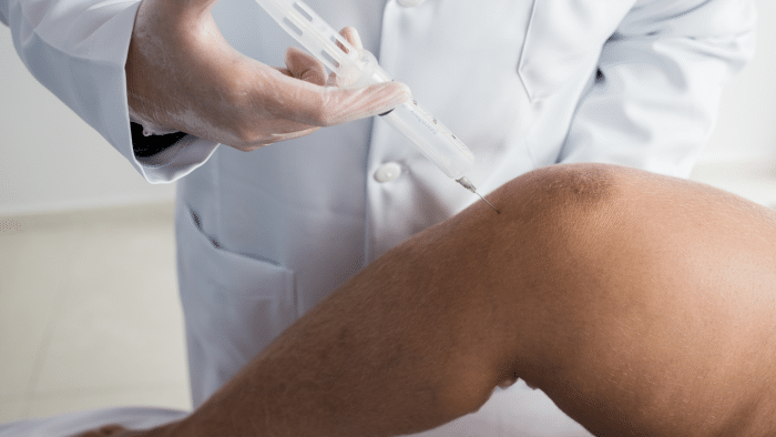 A healthcare professional wearing a white lab coat and gloves administers an injection into a patient's knee, possibly comparing treatments such as Hyalgan vs Euflexxa. The patient's leg is bent, showcasing the knee area where the needle is inserted. The background is a clinical setting.