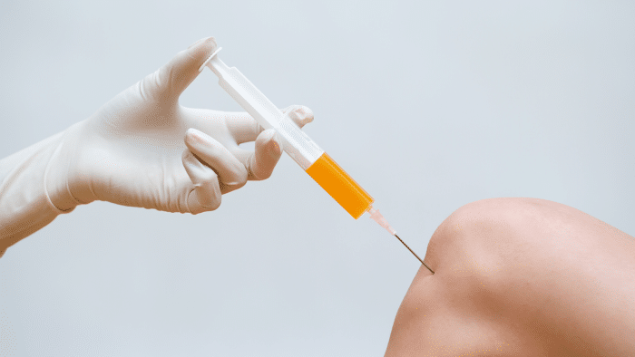 A gloved hand holds a syringe filled with yellow liquid, poised to inject into a person's knee. The background is off-white and out of focus. The scene suggests a medical treatment or procedure involving the knee, specifically the administration of an Orthovisc dosage.