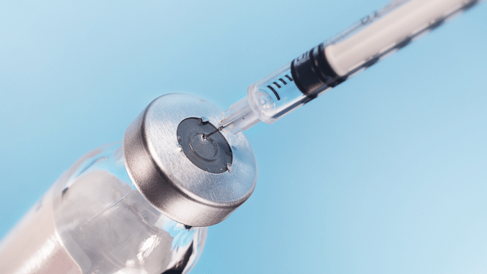 A close-up image of a syringe with the needle inserted into the top of a clear glass vial, ready to draw liquid from it. The background is a solid light blue. The image suggests preparation for an injection or vaccination.