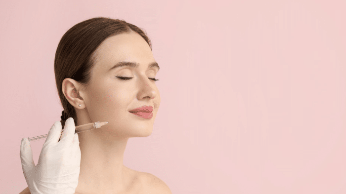 A woman with closed eyes and a serene expression sits against a pink background. A gloved hand holds a syringe near her cheek, indicating a cosmetic procedure or treatment. The woman has light skin, brown hair pulled back, and is wearing minimal makeup.