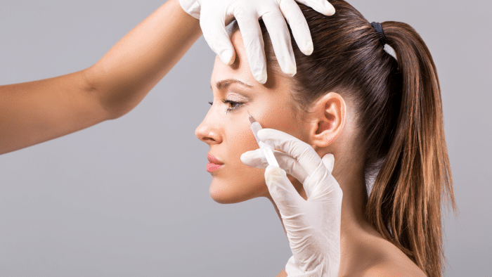 A young woman with her hair in a ponytail is receiving a cosmetic injection on the side of her face from a professional wearing white gloves. Her expression is calm and focused. The background is neutral gray.