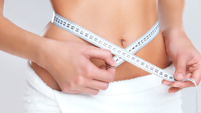 A person measures their waist with a tape measure. The person is wearing a white towel wrapped around their lower body. The measurement is being taken at the midsection, shown from the front torso.