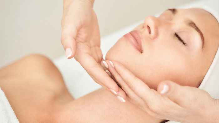 A woman with closed eyes and a serene expression receives a facial massage. Hands are gently placed under her chin, lifting her head slightly. She is wearing a white headband, and the background is softly blurred.