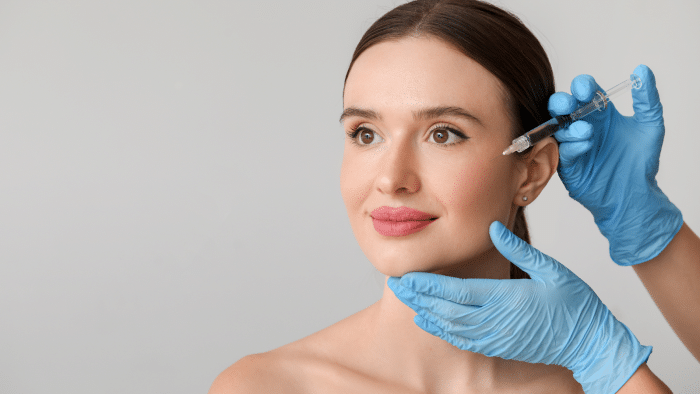 A woman with bare shoulders looks upward and smiles while a pair of gloved hands hold a syringe near her face, preparing for an injection. The background is plain and neutral, highlighting the focus on the cosmetic procedure.