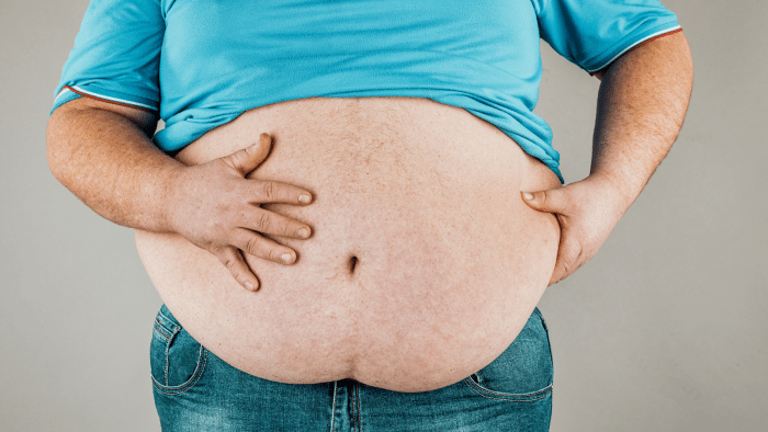 A person wearing a light blue shirt and jeans is shown from the chest down, exposing a large belly. One hand is placed on the hip while the other is gripping the side of the belly, pressing the skin slightly. The background is plain, light gray.