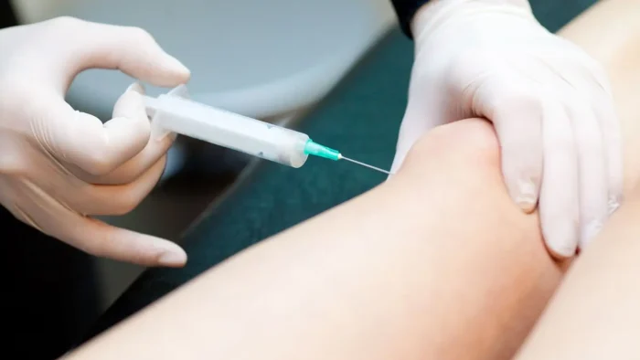 A healthcare professional wearing gloves administers an injection into a patient's knee. The syringe is held in one hand, while the other hand supports the knee. The setting appears to be a medical or clinical environment.