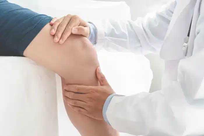 A doctor examines a patient's knee by gently pressing and moving it with their hands. The patient is seated on an examination table, wearing blue shorts. The doctor's white coat and careful attention suggest a medical consultation.