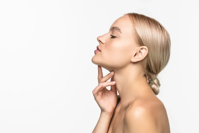 A young woman with blonde hair in a low bun is shown in profile against a white background, eyes closed, gently touching her neck with her hand.