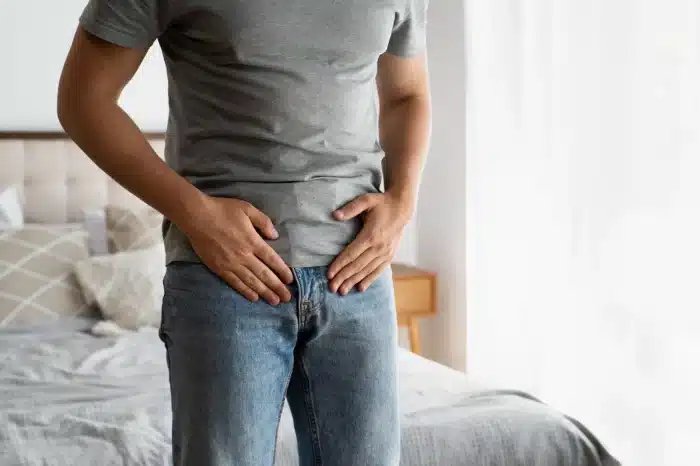A man in a grey t-shirt and jeans stands by a bed, holding his lower abdomen with both hands, appearing to be in discomfort or pain. The room is softly lit with natural light from a window.