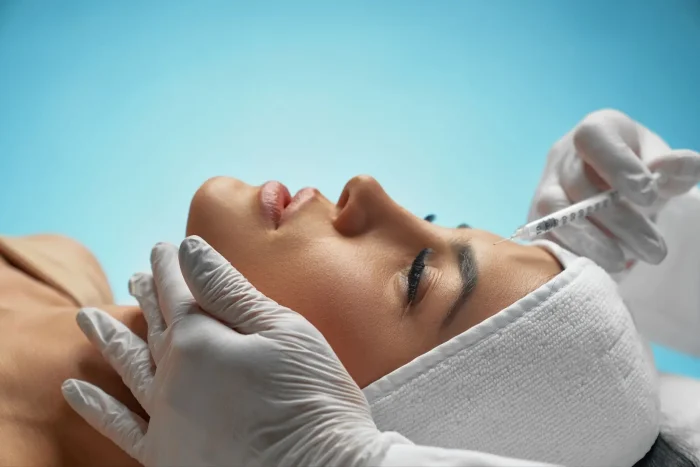 A person with a white headband lies down with eyes closed as a gloved hand injects a syringe into their forehead against a blue background.