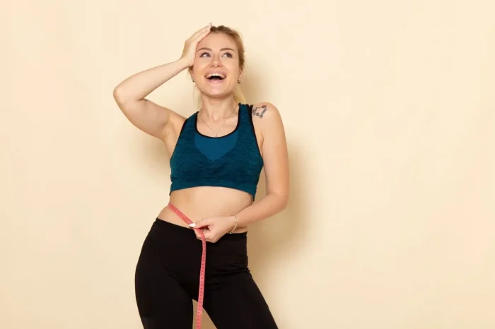 A smiling woman in athletic wear measures her waist with a tape measure, holding one hand to her head, standing against a beige background.